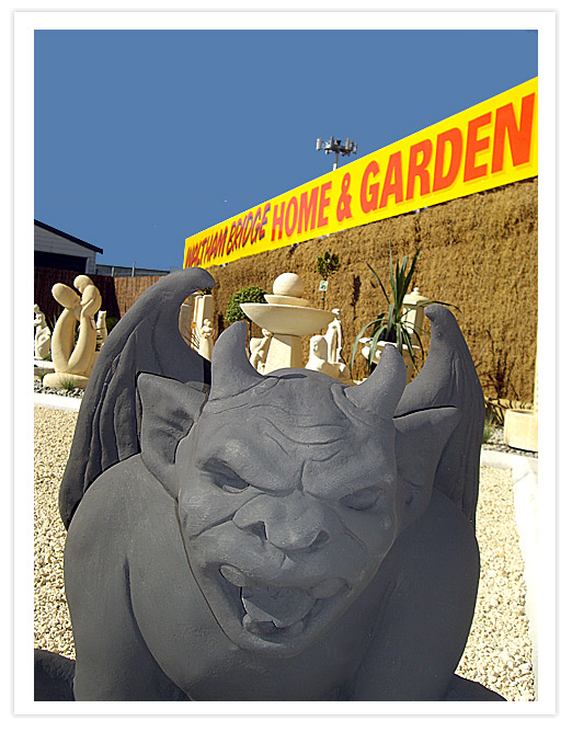 A close-up of a stone gargoyle with wings, positioned in a garden center with a bright yellow sign reading 'Waltham Bridge Home & Garden' in the background.