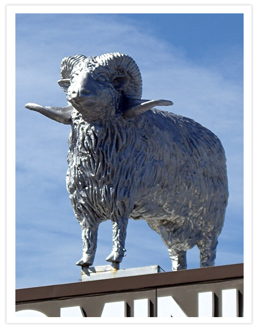 A large silver sculpture of a ram standing on top of the Dominion Carpets building under a blue sky.