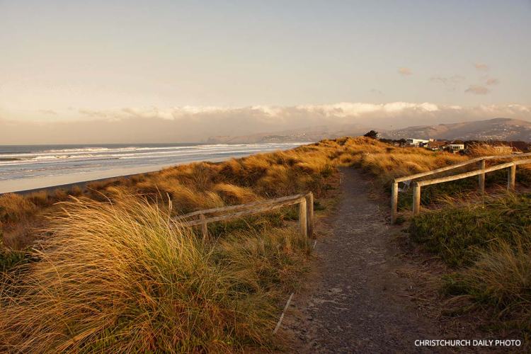 A path leading through tall grass to a beach at New Brighton during late afternoon, with soft waves and distant hills in the background.