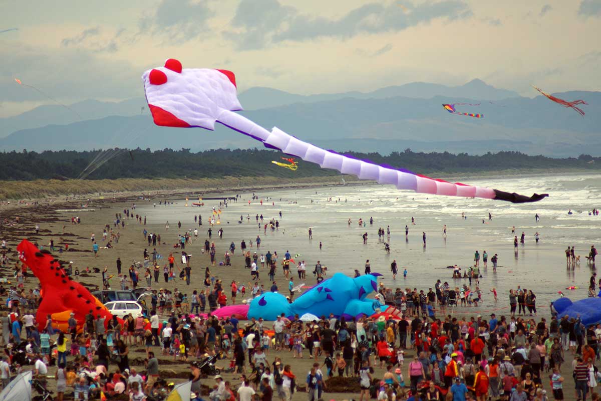 A large crowd enjoying Kite Day on the beach, with colorful kites flying in the sky and inflatable figures on the sand.
