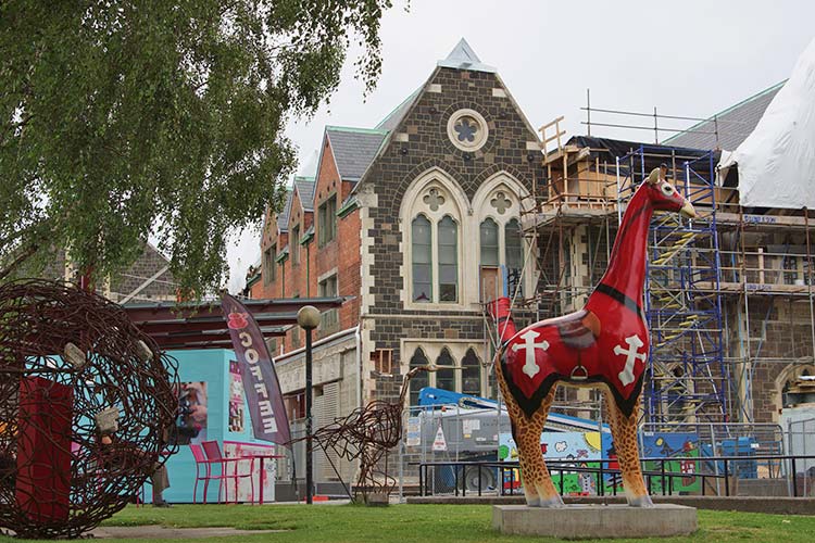 A brightly painted giraffe sculpture stands in front of a historic building undergoing renovation, surrounded by art installations and colorful signs, as part of the Christchurch Stand Tall campaign by artist Martyn Giles.