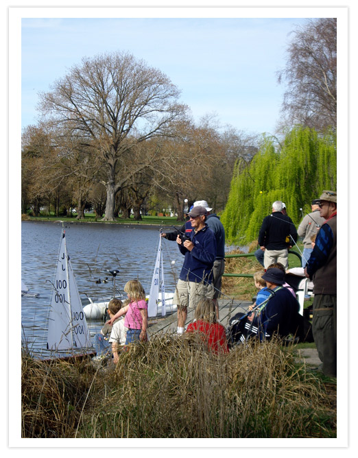 A group of people gathered by a lake, observing small sailboats on the water. There are children playing near the edge, and adults standing or sitting on the shore, surrounded by trees in a park setting.