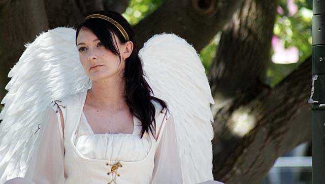 A performer dressed as an angel with large white wings, sitting amidst trees at a festival.