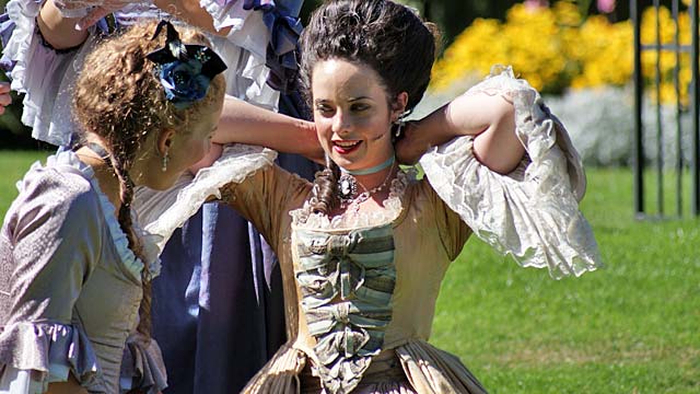 Two actresses in period costumes preparing for a performance outdoors, with a garden background filled with flowers.