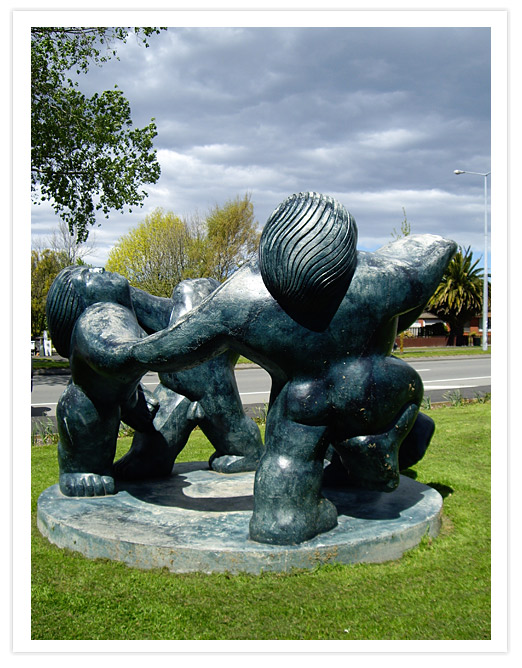 Bronze sculpture titled 'The Joy of Eternal Spring' by Llew Summers, depicting three stylized figures intertwined on a circular base, set against a backdrop of trees and a cloudy sky.