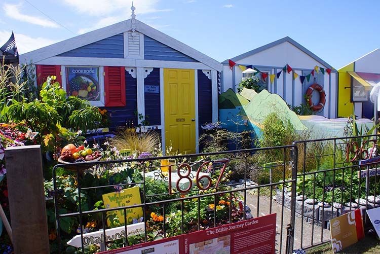 A vibrant garden display featuring a colorful house with a yellow door, surrounded by various plants and flowers at the Ellerslie Flower Show.