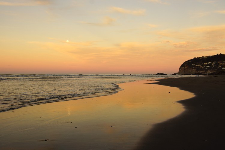 A tranquil beach scene at sunset with soft waves lapping at the shore, reflecting the pastel sky and a distant cliff on the right.