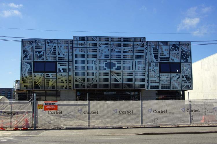 Facade of a modern building on Lichfield Street, featuring an intricate laser-cut steel design resembling the Avon River Loop, with construction barriers in front.