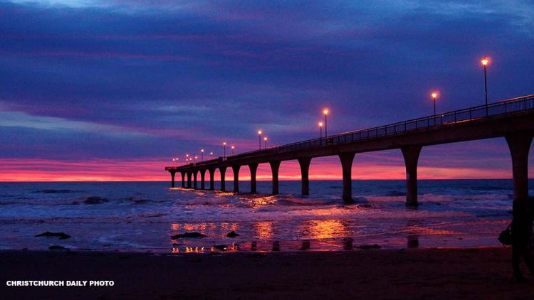 A scenic view of a pier extending over the ocean during sunset, with lights illuminating the structure and reflecting on the water.