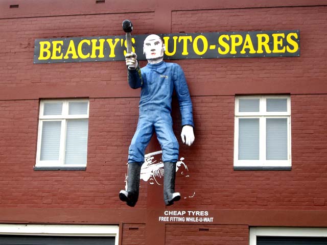 A large, painted figure of a mechanic in blue overalls hanging outside a store called Beachy's Auto-Spares, with a sign advertising cheap tyres and free fitting.
