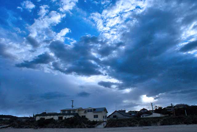 A dramatic evening sky over Kaka Point beach with dark clouds and a hint of blue, showcasing nearby houses amid stormy weather.