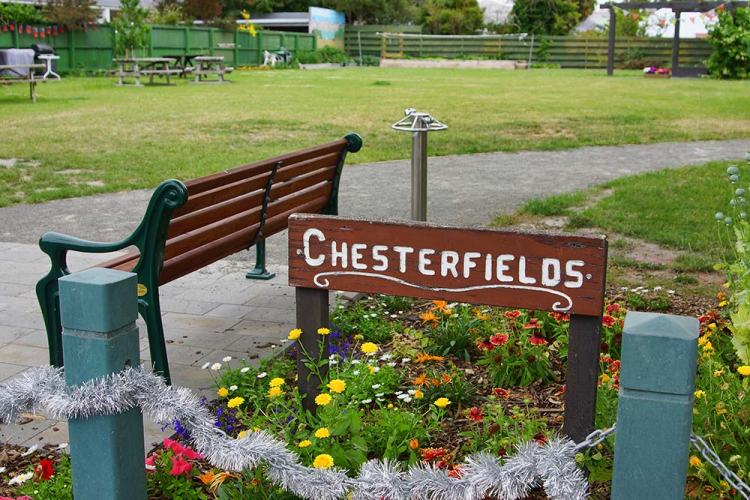 A sign reading 'Chesterfields' in a community park with a wooden bench and colorful flower beds.