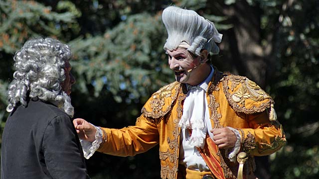 Two actors in period costumes performing a scene outdoors, surrounded by trees.