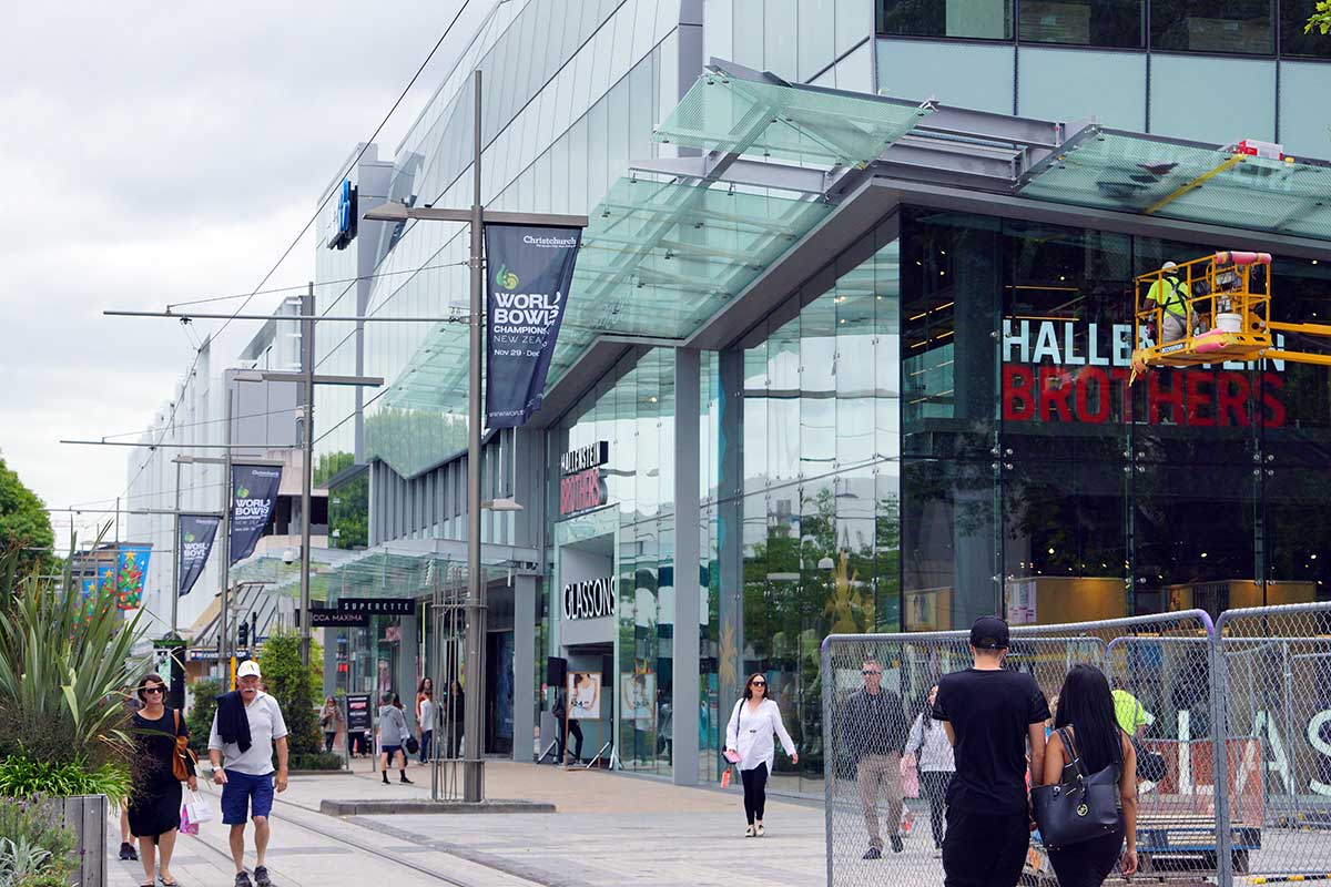 View of the bustling Cashel Street in Christchurch featuring the newly opened ANZ Centre, with pedestrians walking by shops including Hallenstein Brothers and Glassons. Construction work is also visible in the area.