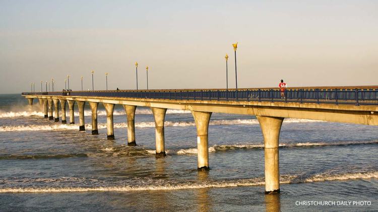 A long pier extends into the ocean with a person walking along it, surrounded by waves and a clear sky.