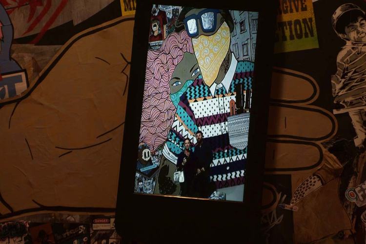 Interactive street art display featuring colorful artwork of two figures. One figure has pink hair and is wearing a patterned sweater, while the other is dressed in glasses and a patterned shirt. A couple is posing in front of the artwork.