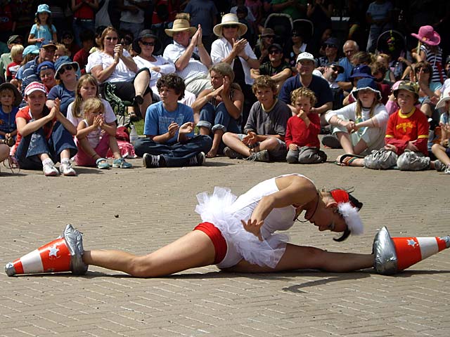 A performer dressed in a tutu and festive accessories is executing a split on the ground, flanked by traffic cones, in front of an audience at the Christchurch Busker's Festival.