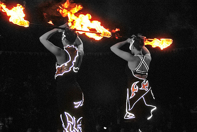 Two performers in black and white with flames, dressed in illuminated costumes, showcasing a fire-dancing performance.