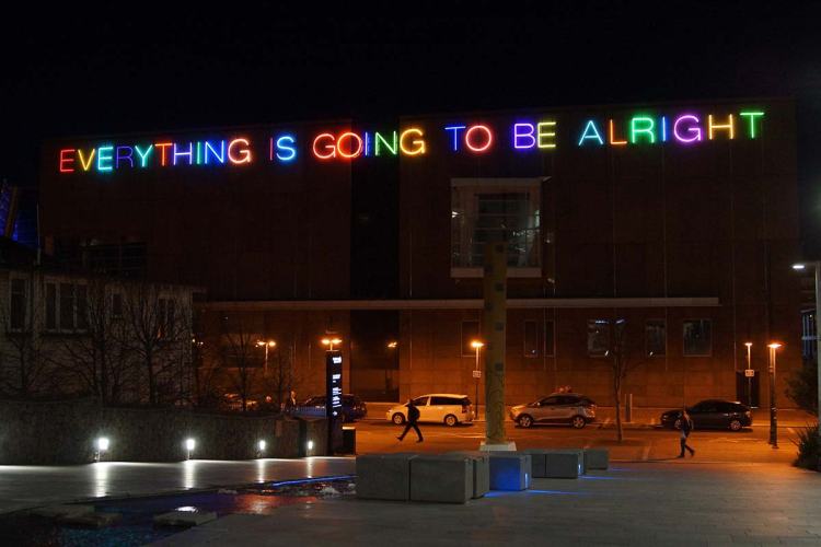 A brightly lit neon sign on the exterior of the Christchurch Art Gallery reads 'EVERYTHING IS GOING TO BE ALRIGHT' at night, showcasing vibrant colors against the building.
