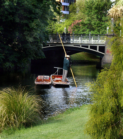 A punter stands on a small boat, using a pole to navigate through a calm waterway beneath a decorative bridge, surrounded by lush greenery.