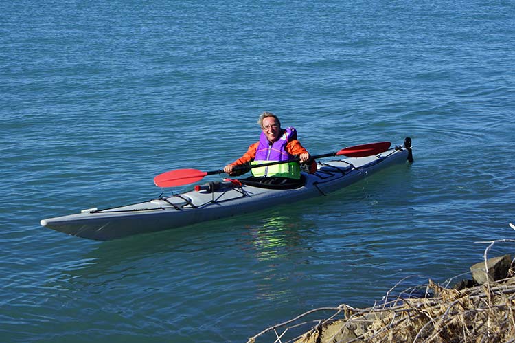 A person in a kayak wearing a colorful life jacket paddles in a calm estuary water.