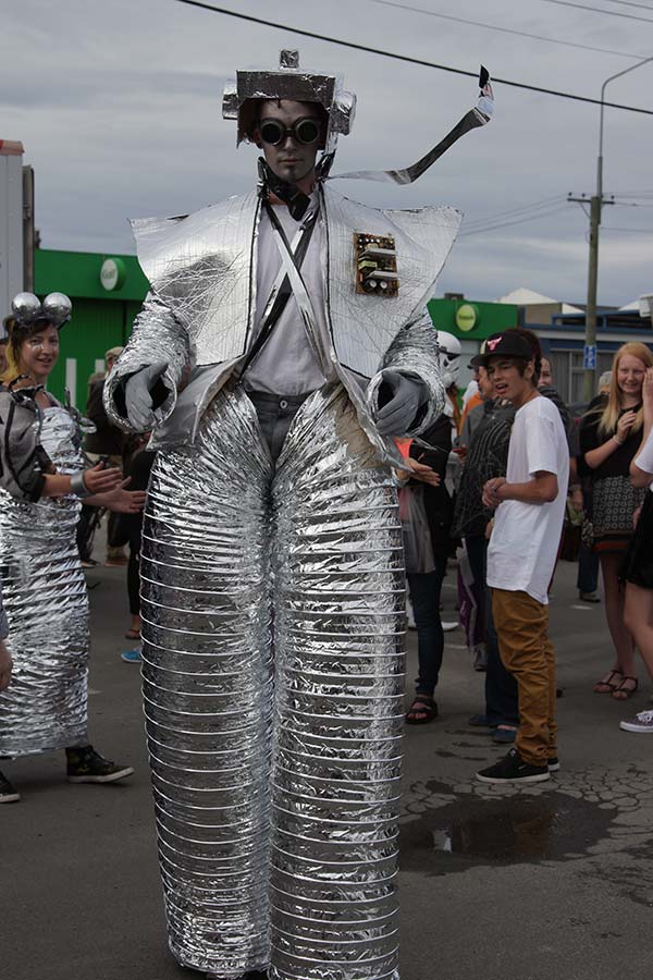 A performer in a large silver robot costume made of metallic material, standing on stilts, at a street event with an audience in the background.