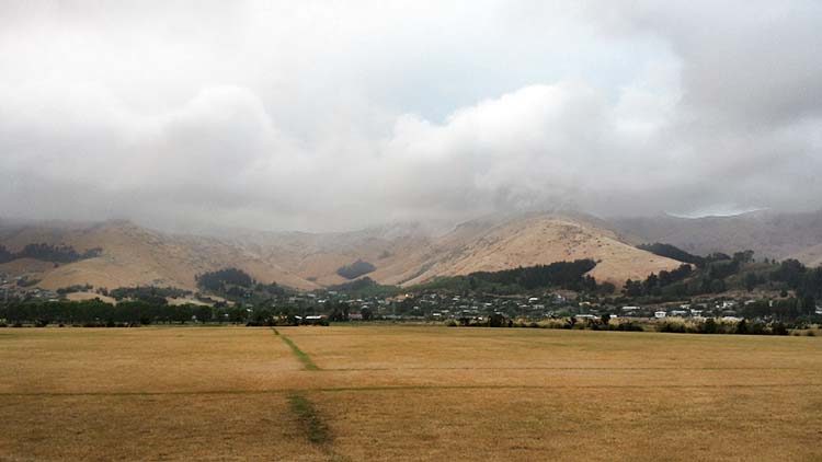 A panoramic view of hills covered in low clouds, with a foreground of dry grass and a small town visible in the valley.