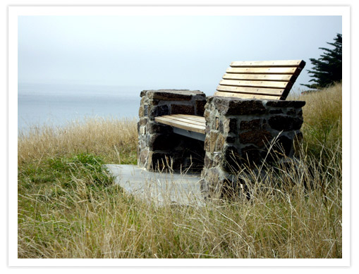 A stone and wooden bench overlooking Taylor's Mistake beach, surrounded by tall grasses and a scenic view of the ocean.
