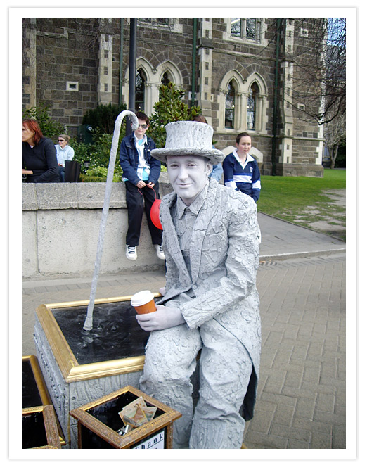 A living statue in a gray suit and top hat holding a coffee cup, seated next to a donation box at the Christchurch Arts Centre, with people in the background.
