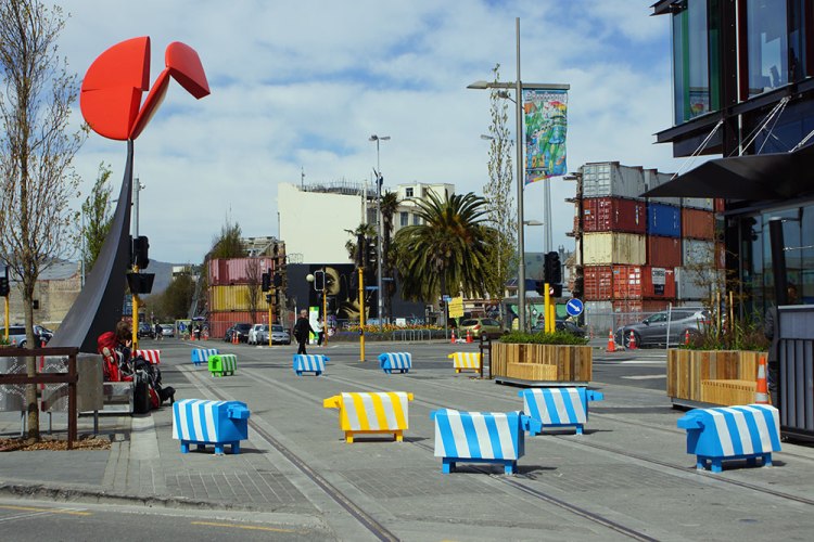 A flock of colorful striped plastic sheep positioned on a city street, with modern buildings and palm trees in the background.