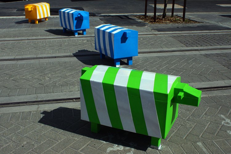 A flock of colorful, striped plastic sheep in yellow, blue, and green arranged on a cobblestone street, adding a whimsical touch to the environment.