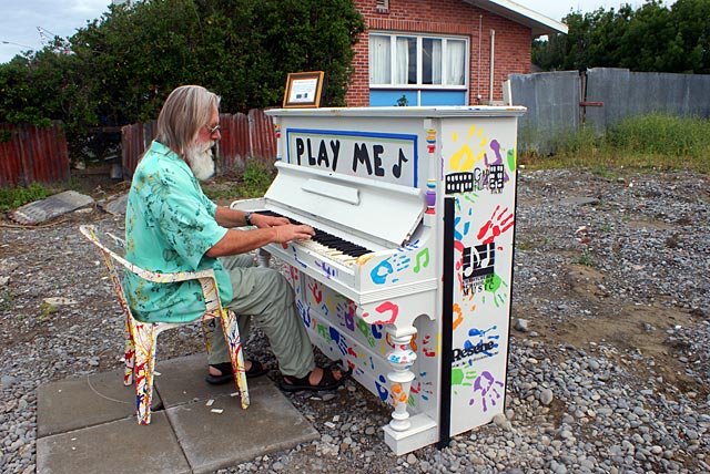 A man with long hair and a beard plays a colorful, decorated piano in an empty lot. The piano has the words 'PLAY ME' on the front, with various artistic designs around it.
