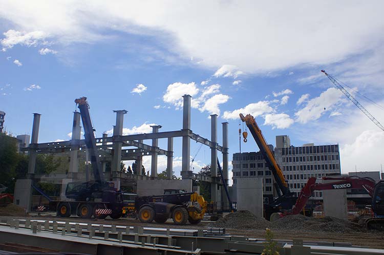 Construction site with cranes and machinery, showcasing the framework of a building against a blue sky with clouds.