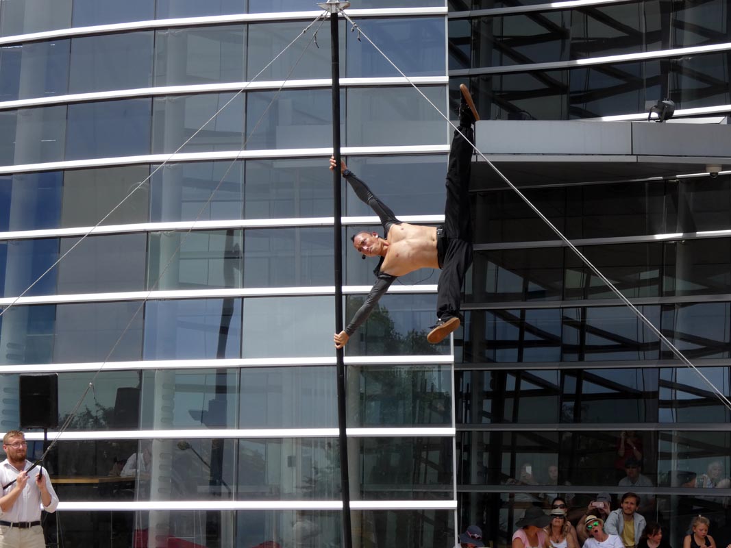 A performer hanging from a vertical pole outside an art gallery, demonstrating an impressive acrobatic pose, with an audience watching in the background.