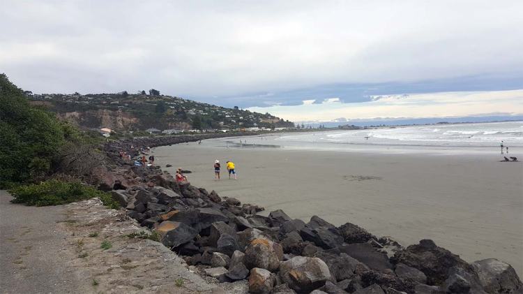 A beach scene with people walking along the shore, rocky formations in the foreground, and a cloudy sky in the background.