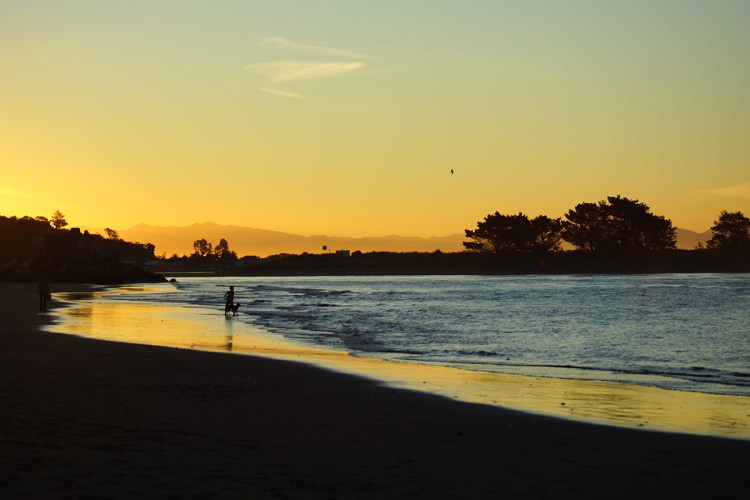 Silhouette of a person walking along Sumner Beach during sunset with the sun setting over the horizon, casting a warm glow on the water and sand.