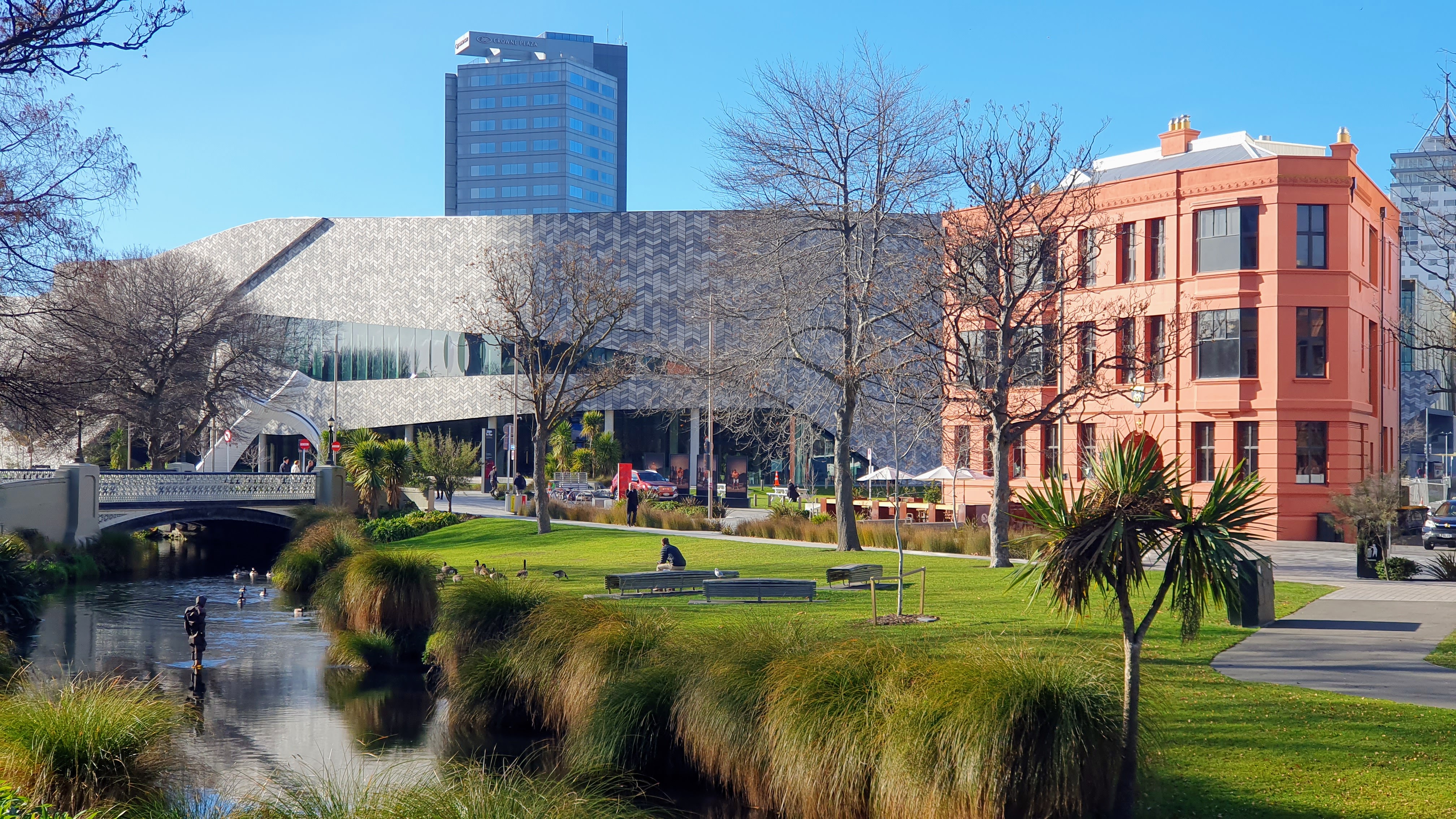 A scenic view featuring a river alongside green grass, native plantings, a sculpture in the river, the Te Pae Convention Centre, and the Midland Club building.