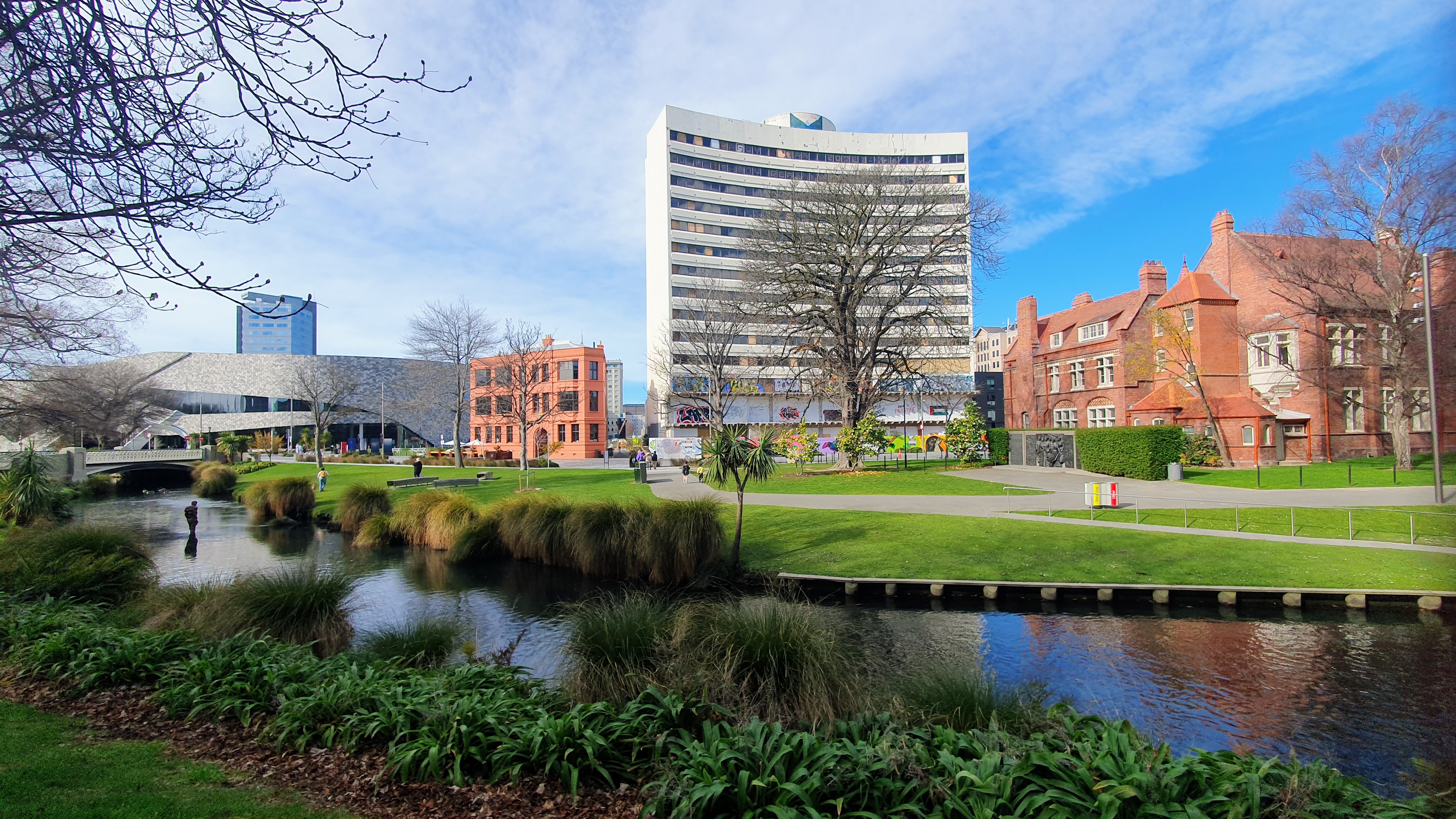 A scenic view of the Avon River with a pedestrian walkway, featuring the Te Pae Convention Centre on the left, the newly restored Municipal Chambers in the center, and the old Noahs/Rydges Hotel in the background.