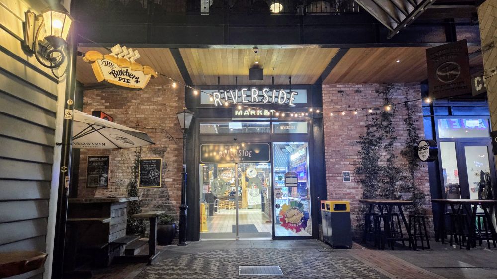 Entrance of Riverside Market, featuring a sign for 'The Butcher's Pie' and outdoor seating with string lights.