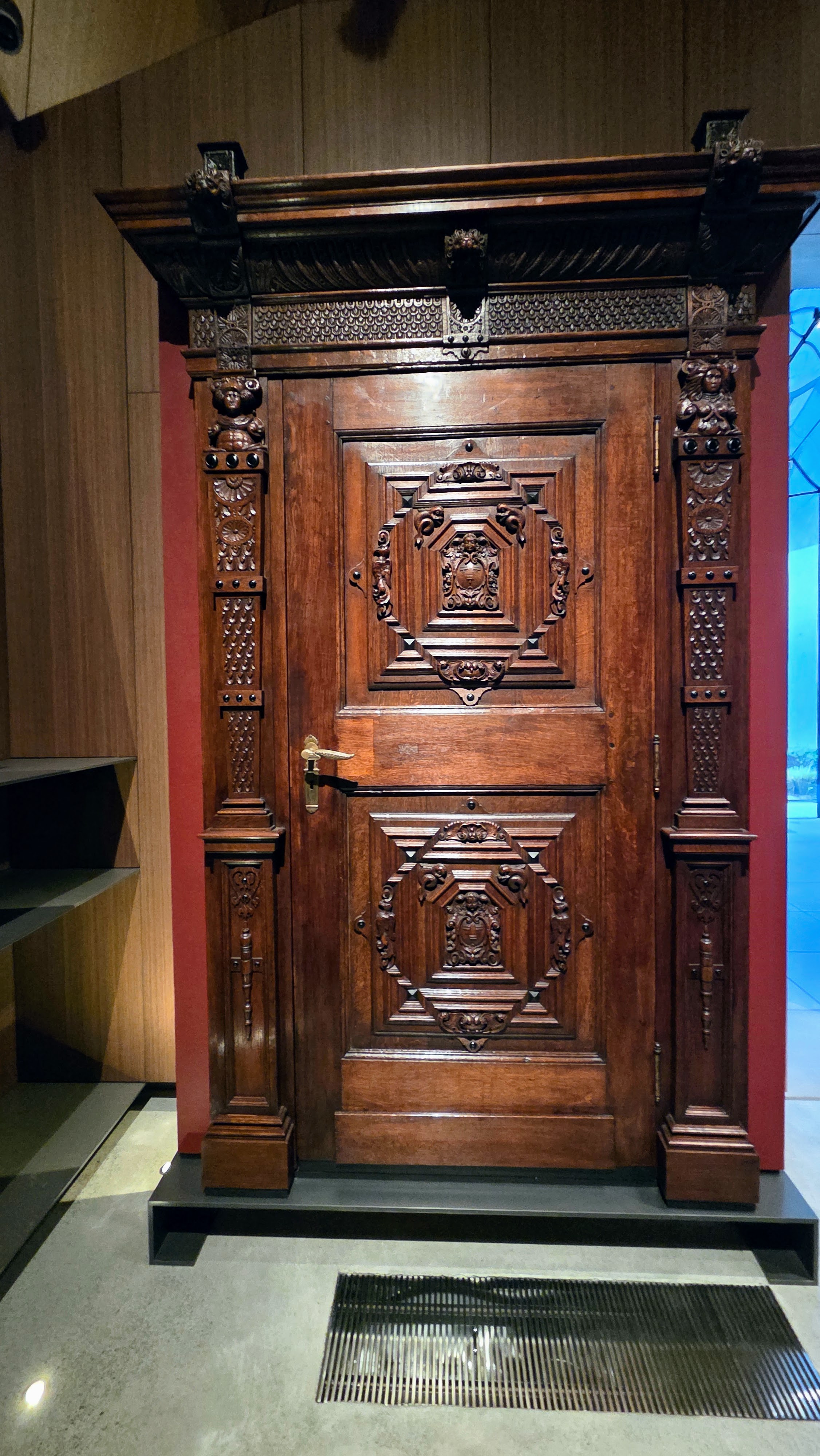 An intricately carved wooden door with ornate detailing, set against a muted background in a museum setting.
