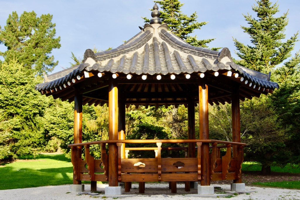 A traditional Korean jeongja pavilion made of wood, featuring a curved roof with tiles, situated in a green garden setting.