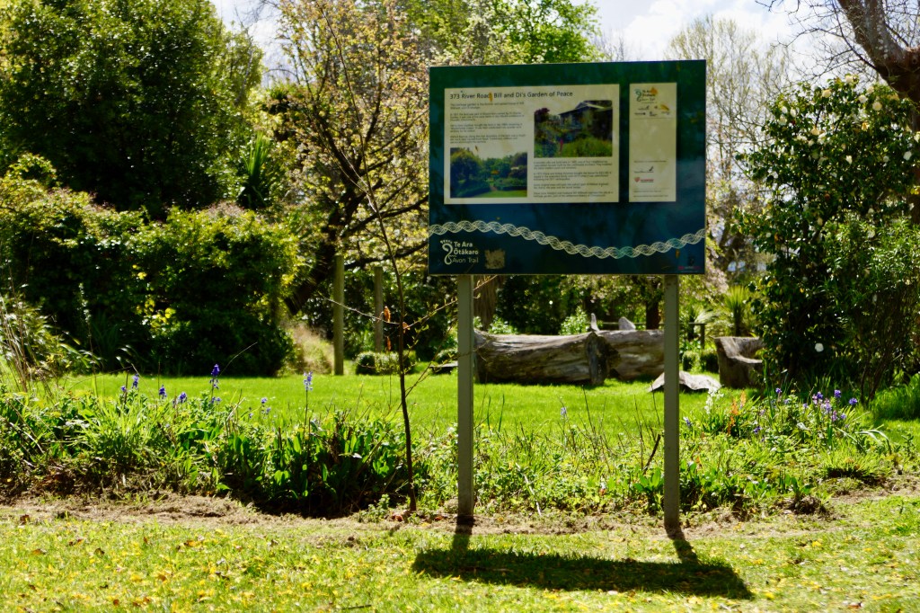 A sign in a garden with text about Bill and Di's Garden at 373 River Road, surrounded by greenery and flowering plants.