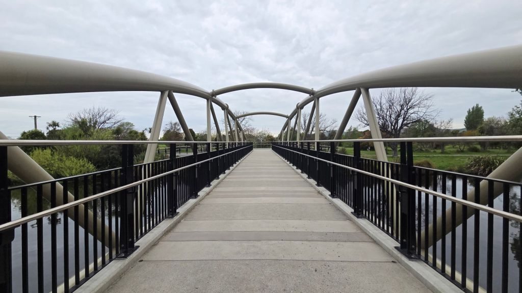 A modern footbridge with metal arches spans over a river, surrounded by greenery under a cloudy sky.