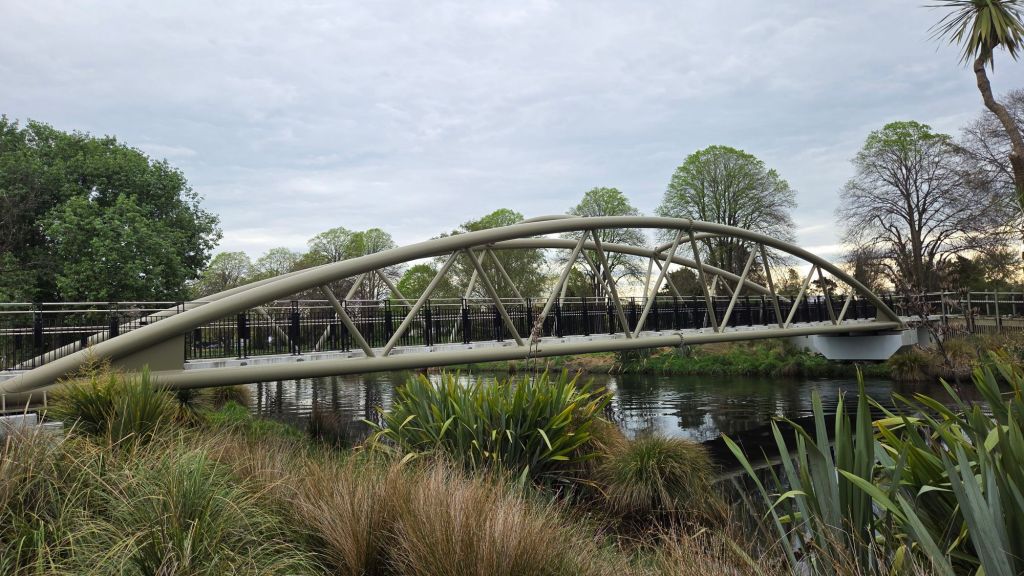 A modern footbridge with a curved design spans over a calm waterway, surrounded by greenery and trees under a cloudy sky.