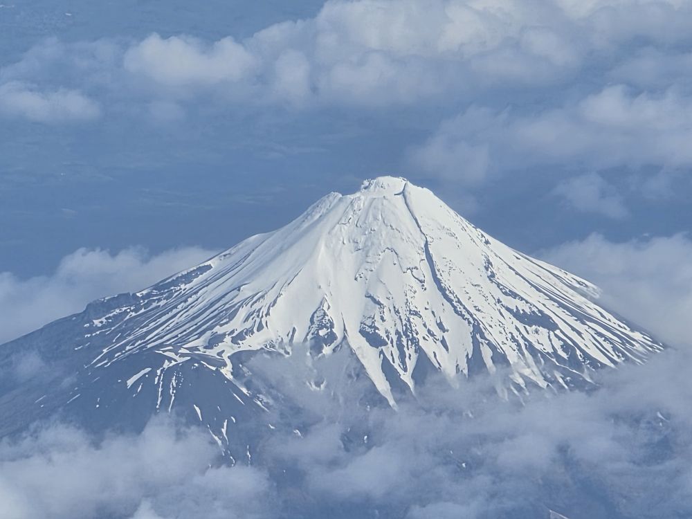 Aerial view of Mt Taranaki, showcasing its snow-capped peak surrounded by a blue sky and wispy clouds.