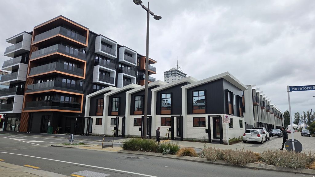 Modern townhouse development in a central city area, featuring dark and light-colored buildings alongside a wide road and green landscaping.