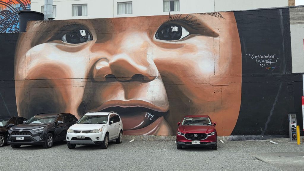 A large-scale mural titled 'Unfinished Infancy' by street artist RichiePopsBaker, depicting a close-up of a baby's face with a playful expression. The mural is located on Manchester Street in central Christchurch, with parked cars in front that seem to complement the artwork.