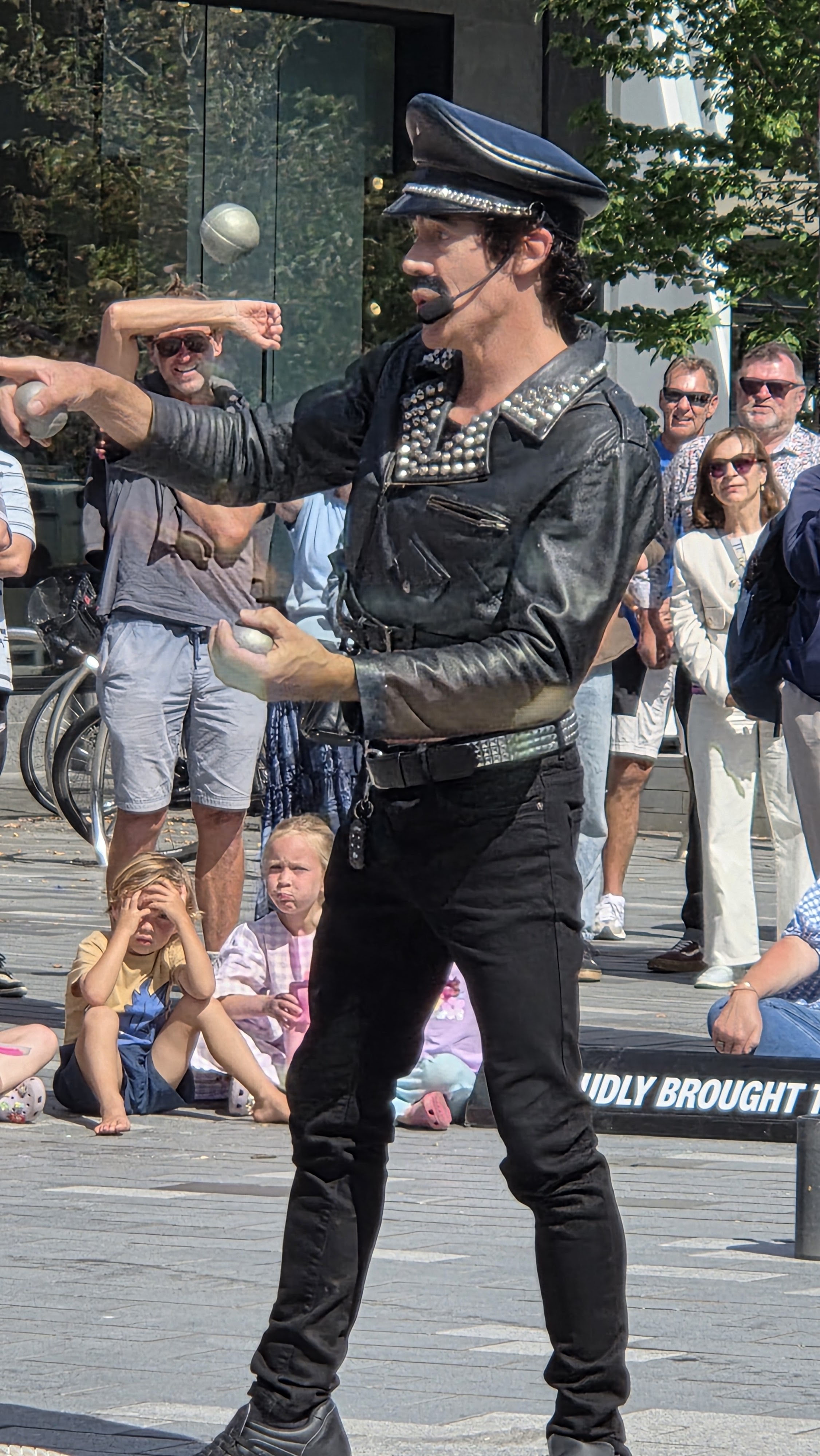 A street performer in a leather jacket and hat juggles while entertaining a crowd gathered in a city square. Spectators, including children, watch intently.