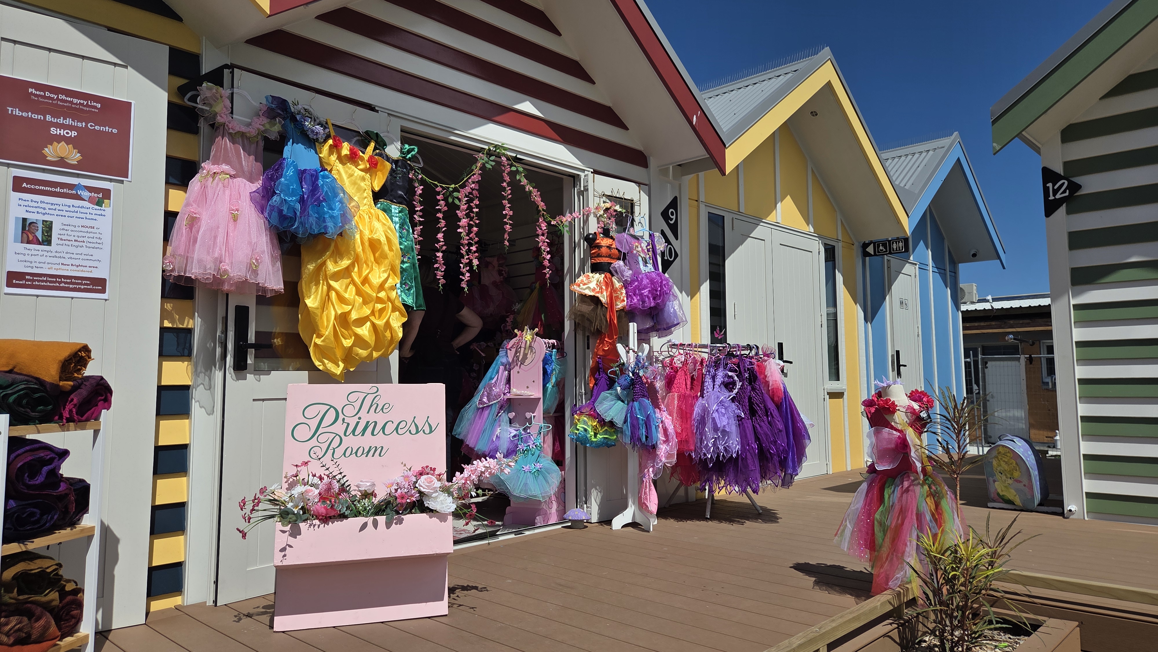 Front of 'The Princess Room' shop featuring colourful costumes hanging outside, including princess dresses and tutus. A sign displays the shop's name, surrounded by decorative flowers. In the background, vibrant beach hut-style buildings are visible under a clear blue sky.